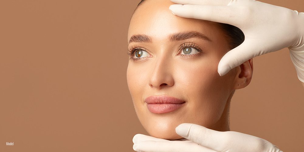 two hands in white surgical gloves frame a woman's face as she looks off into the distance